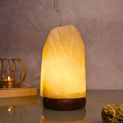 Glowing Himalayan salt lamp with wooden base on reflective surface, candle and sage bundle nearby