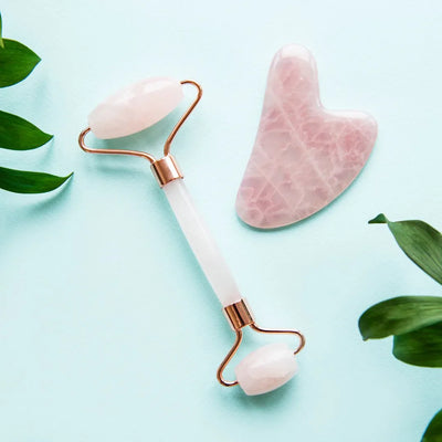 Rose quartz facial roller and gua sha tool on light blue background with green leaves