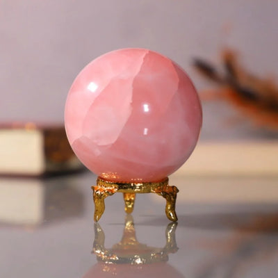 Polished pink rose quartz crystal sphere on ornate gold stand with blurred background